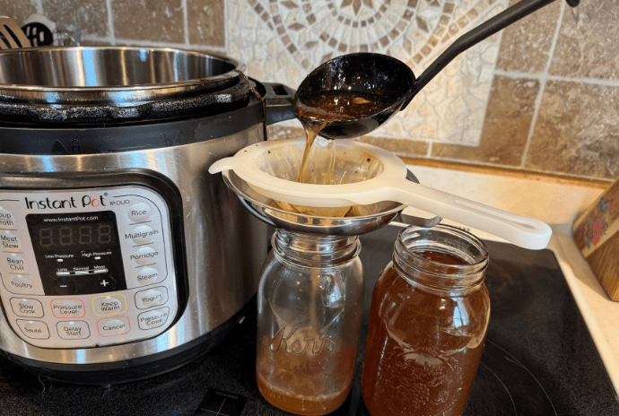 Ladling and straining hot beef bone broth into a mason jar using a funnel and fine mesh strainer