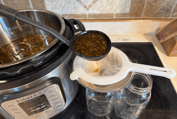 Ladling and straining hot beef bone broth into a mason jar using a funnel and fine mesh strainer