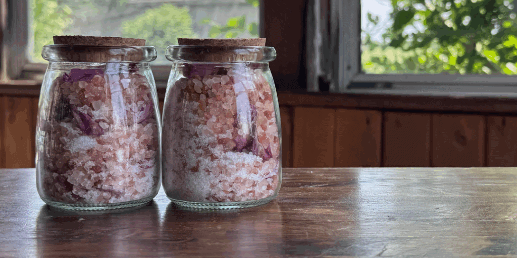 Glass jars filled with herbal bath salts and flower petals on a rustic surface.