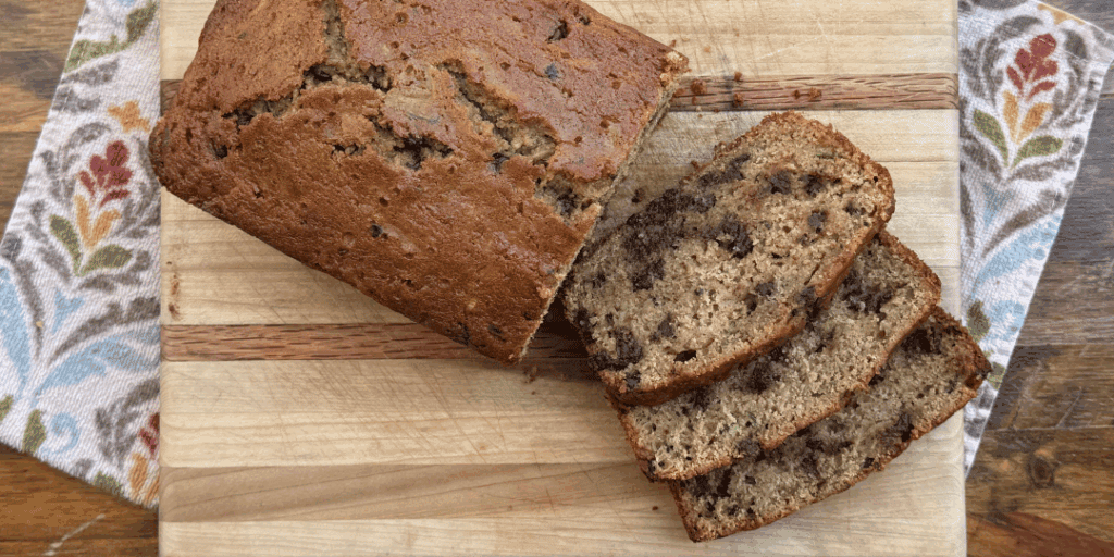 sliced chocolate chip zucchini bread on a wood cutting board, kitchen towel, and wooden table