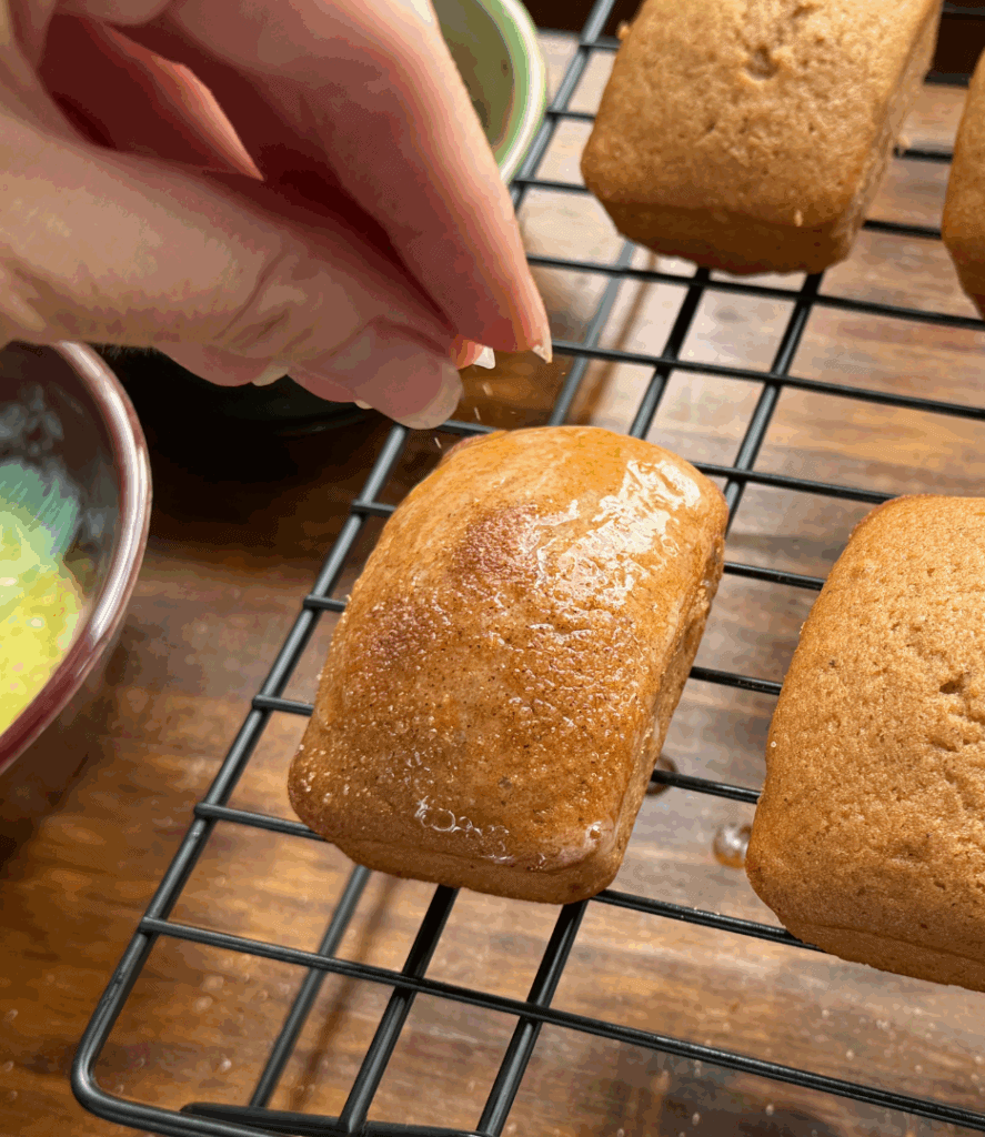 sprinkling cinnamon sugar on homemade apple cider donut mini bread loaves on a cooling rack